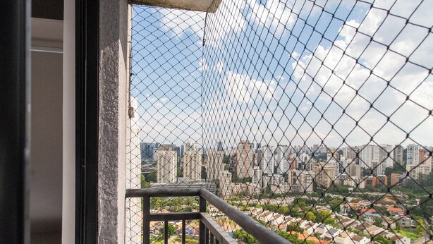 A balcony with a view of a cityscape, featuring a chain-link fence and a black railing. - Residencial Pacobá (São Paulo)