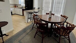 Uma sala de jantar com mesa redonda e cadeiras de madeira, cozinha ao fundo e uma janela com persianas.