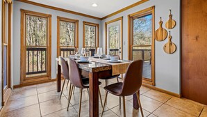 Comedor con mesa de madera dispuesta para cuatro personas, sillas, ventanas amplias con vista a árboles y decoración de pared de madera.
