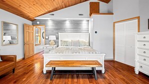 Un dormitorio con techo de madera, una cama grande con cabecera blanca, un banco de madera, un aparador blanco y un armario.