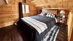 A wooden cabin bedroom with a bed, a nightstand, a lamp, and a window. - Black Bear, Black Bear Lakefront Cabin (Cantwell)