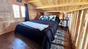 Een slaapkamer in een houten blokhut, met een bed, nachtkastje, ventilator en een raam.