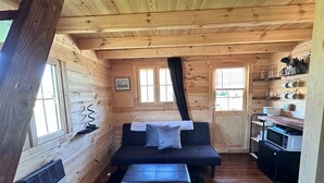 Interieur van een blokhut met een bank, een kleine tafel, een keukenhoek en een raam met gordijnen.