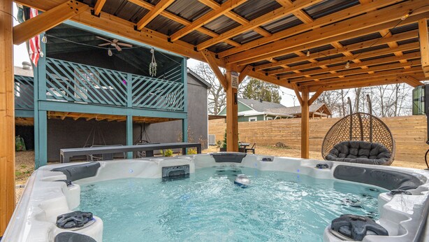 Un espace avec un jacuzzi, surmonté d’une pergola en bois, un coin salon abrité et une vue sur une maison au loin.