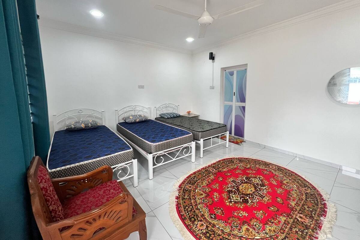 Une pièce avec deux lits à ossature métallique, une chaise en bois, un tapis rouge à motifs, un ventilateur de plafond et une porte aux motifs bleus et blancs.