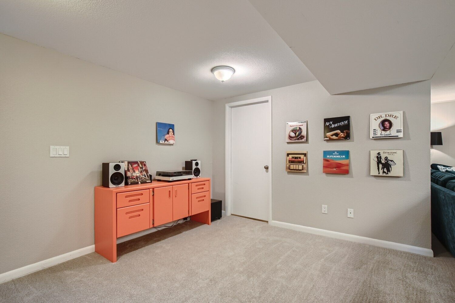 A room with a beige carpet, a white door, and a white ceiling. There is a small orange cabinet with speakers and a framed picture on the wall.