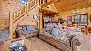 Ein gemütliches Blockhaus-Interieur mit einer Holztreppe, einem Wohnbereich mit zwei Sofas, einem Esstisch und einer Küchenzeile.