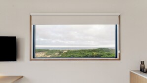 A room with a large window overlooking a natural landscape, a flat-screen TV mounted on the wall, and a wooden bench. - Offshore Montauk (Montauk)