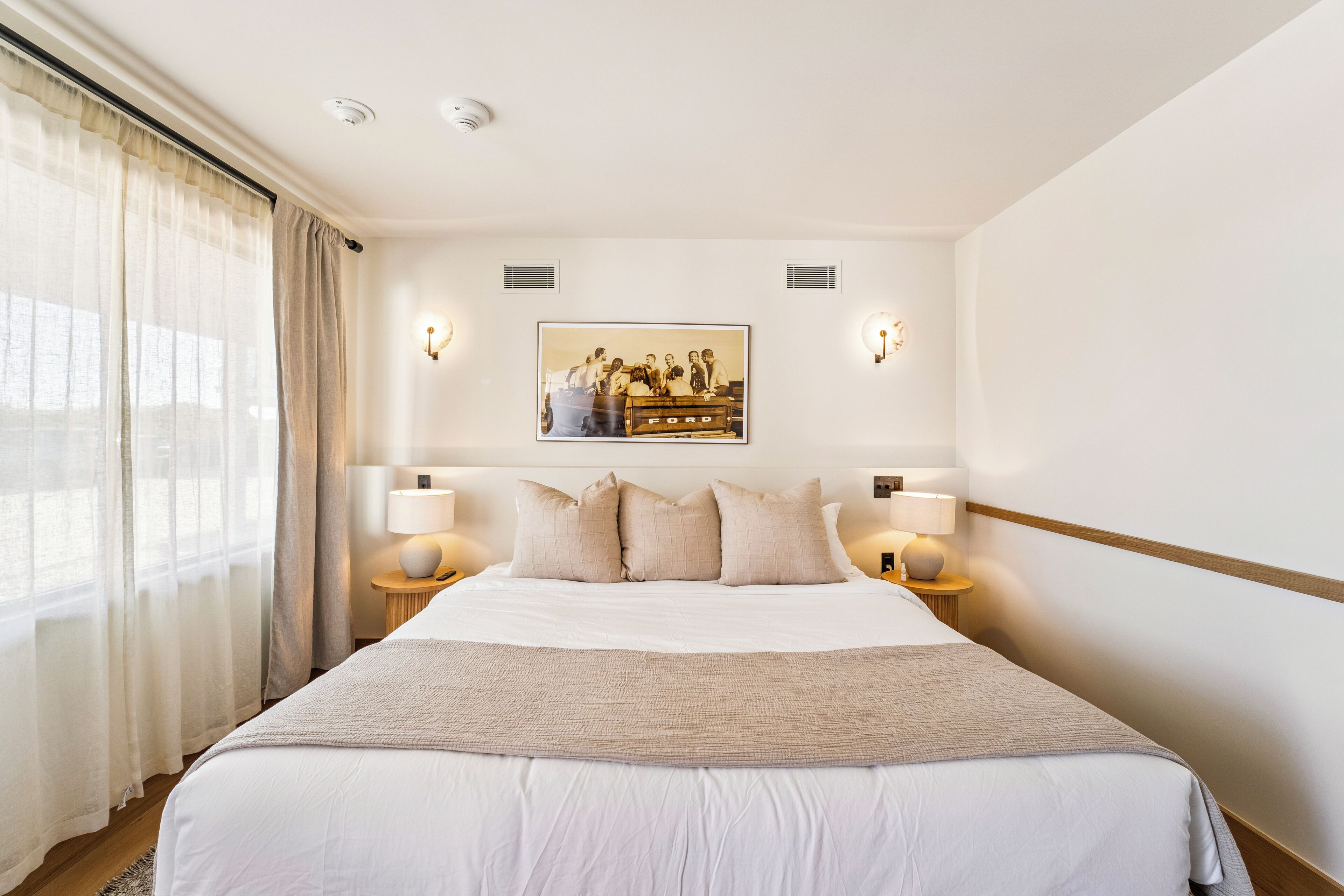 A hotel room with a large bed, two bedside tables with lamps, a framed picture on the wall, and sheer curtains on the window.