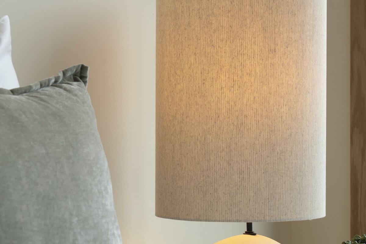 Un lit avec une literie blanche, un oreiller gris et une lampe avec un abat-jour beige.