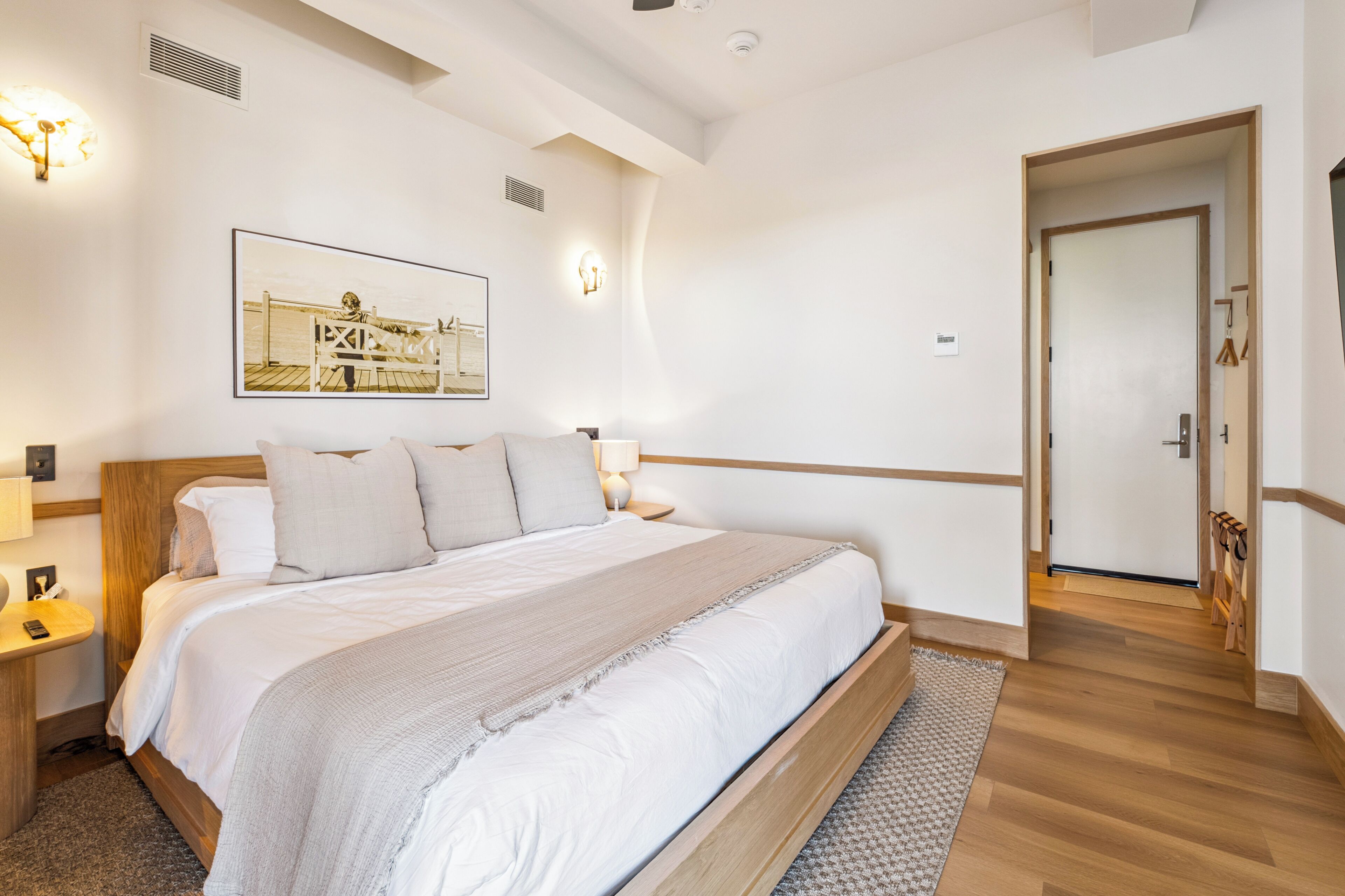 Une chambre à coucher bien rangée, avec un grand lit, une table de chevet, une commode, une lampe fixée au mur et un tableau encadré accroché au mur.