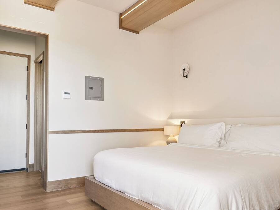A modern hotel room with a wooden floor, a large bed with white bedding, a nightstand, and a wall-mounted light fixture.