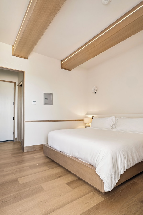 A modern hotel room with a wooden floor, a large bed with white bedding, a nightstand, and a wall-mounted light fixture. - Offshore Montauk (Montauk)