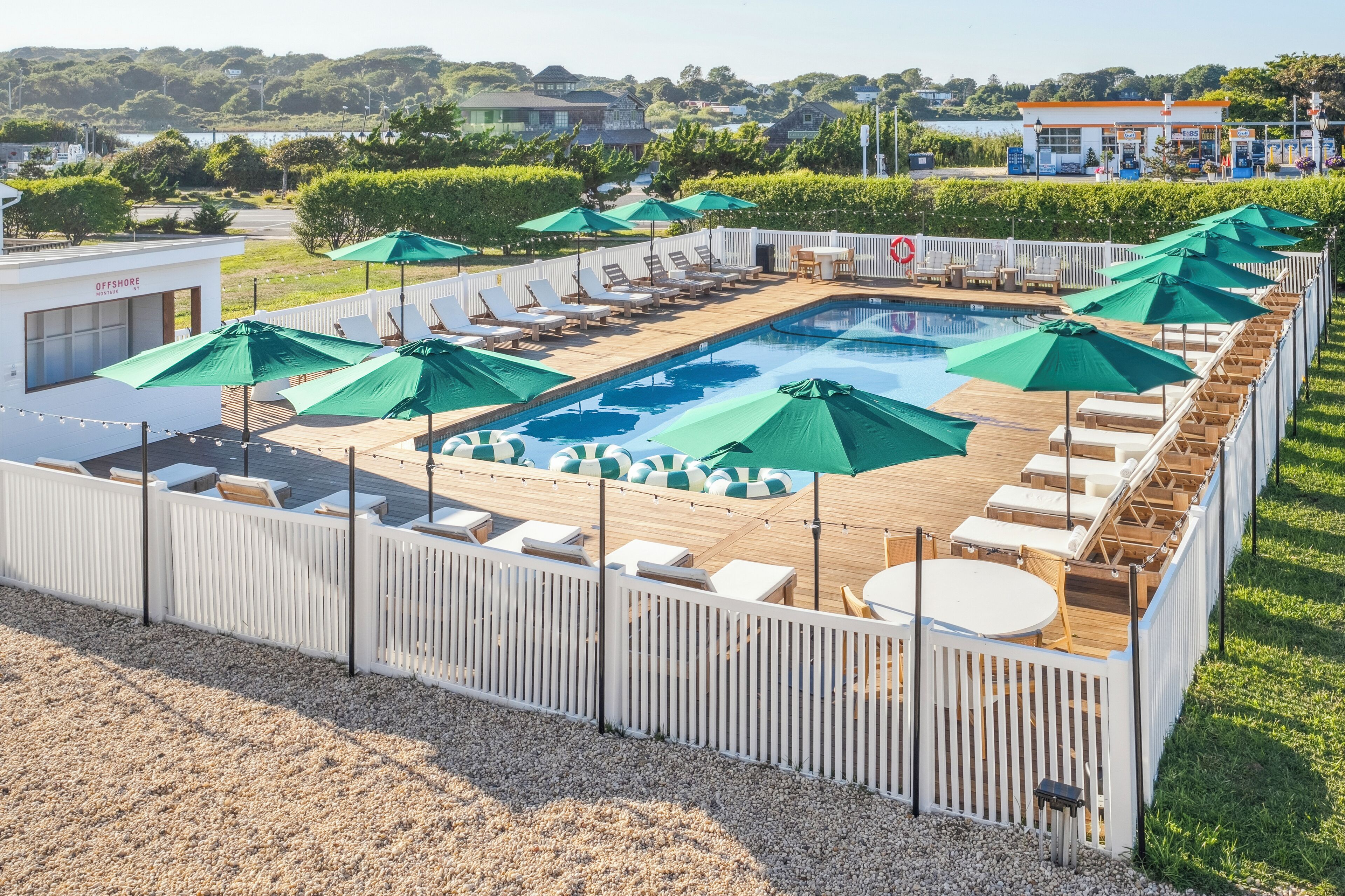 Un espace piscine doté de chaises longues et de parasols, entouré d’une terrasse en bois et d’une clôture blanche.