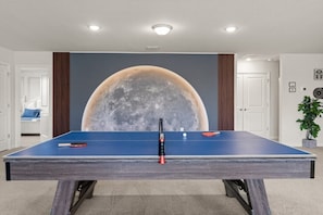Una mesa de ping pong con un gran mural lunar al fondo, una puerta a la izquierda y una planta en maceta a la derecha.