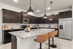 Una cocina moderna con armarios de madera oscura, encimera de granito, electrodomésticos de acero inoxidable, iluminación colgante y una isla central con taburetes de bar.