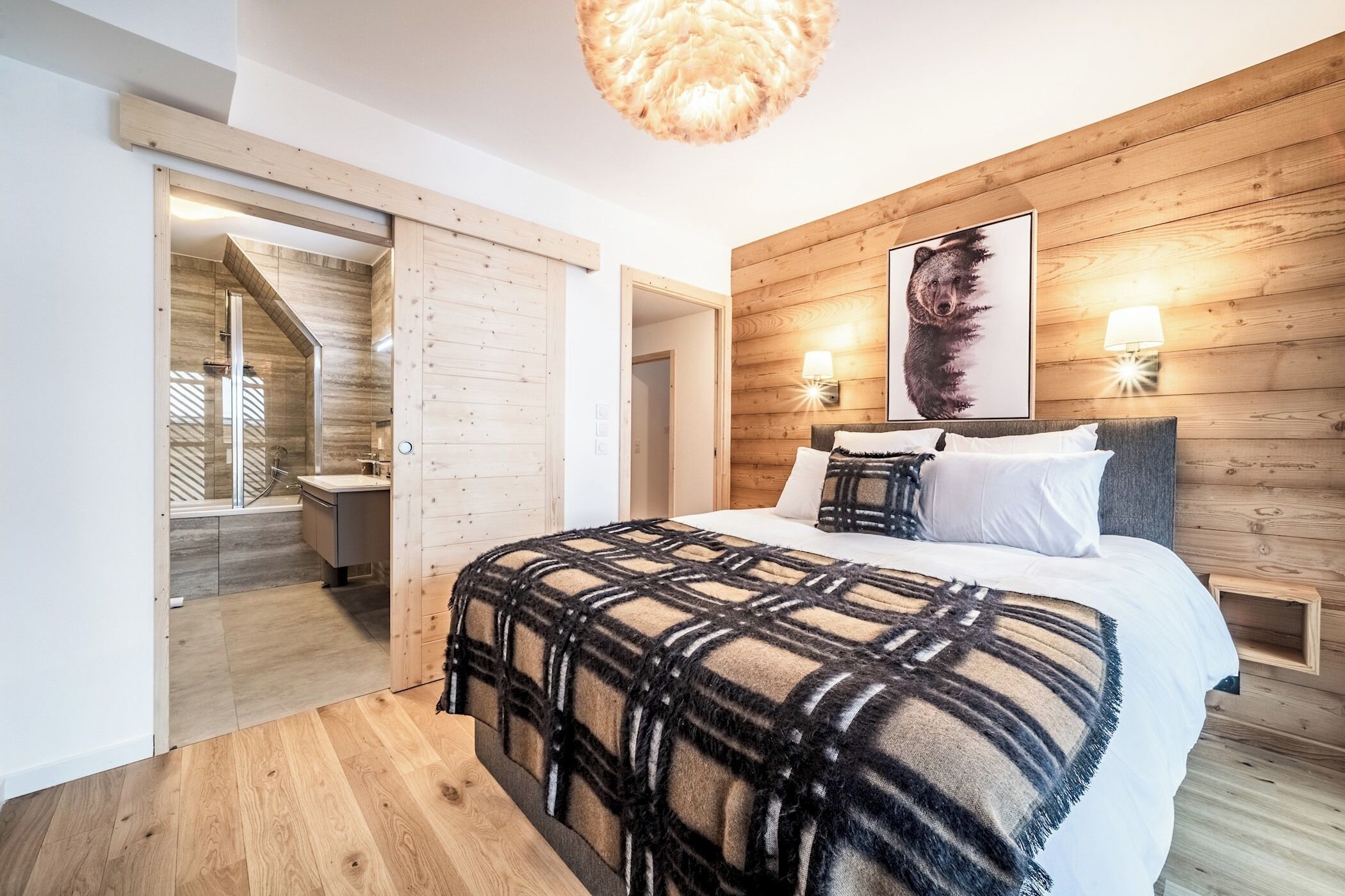 Ein modernes Schlafzimmer mit einem Holzkopfteil, einem großen Bett mit einer karierten Decke, einer Wandleuchte und einem Bären-Motiv-Kunstwerk.