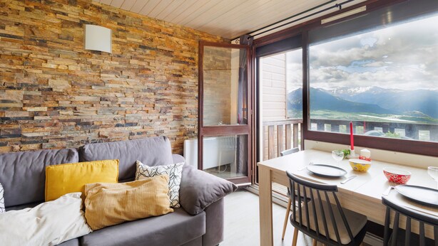 A room with a stone wall, a grey sofa with cushions, a wooden table with a glass, and a view of mountains through a large window. - ANANA Balcon Cerdan | Duplex with Mountain Views (Font-Romeu-Odeillo-Via)