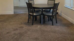 A dining area with a round table and four chairs, a ceiling fan, and a kitchen in the background.