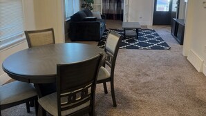 A spacious living room with a dark wood dining table and chairs, a black sofa, and a large window.