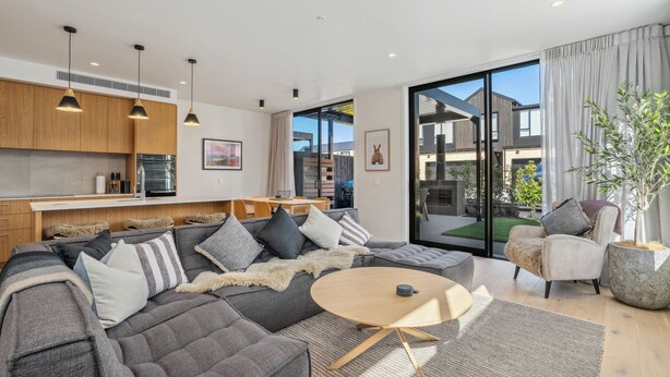 A modern living room with a grey sectional sofa, a round coffee table, and a dining area with wooden cabinets and a kitchen in the background. - Explorers Rest - Wanaka Holiday Home (Wanaka)