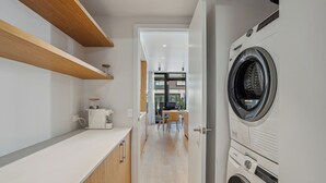 A modern laundry room with a front-loading washing machine, wooden shelves, and a kitchen area in the background. - Explorers Rest - Wanaka Holiday Home (Wanaka)