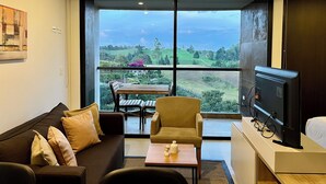 A modern living room with a sofa, armchair, and a small table. A television is on a stand to the right. Large windows offer a view of a green landscape. - Suites - Hotel Rioverde (Rionegro)