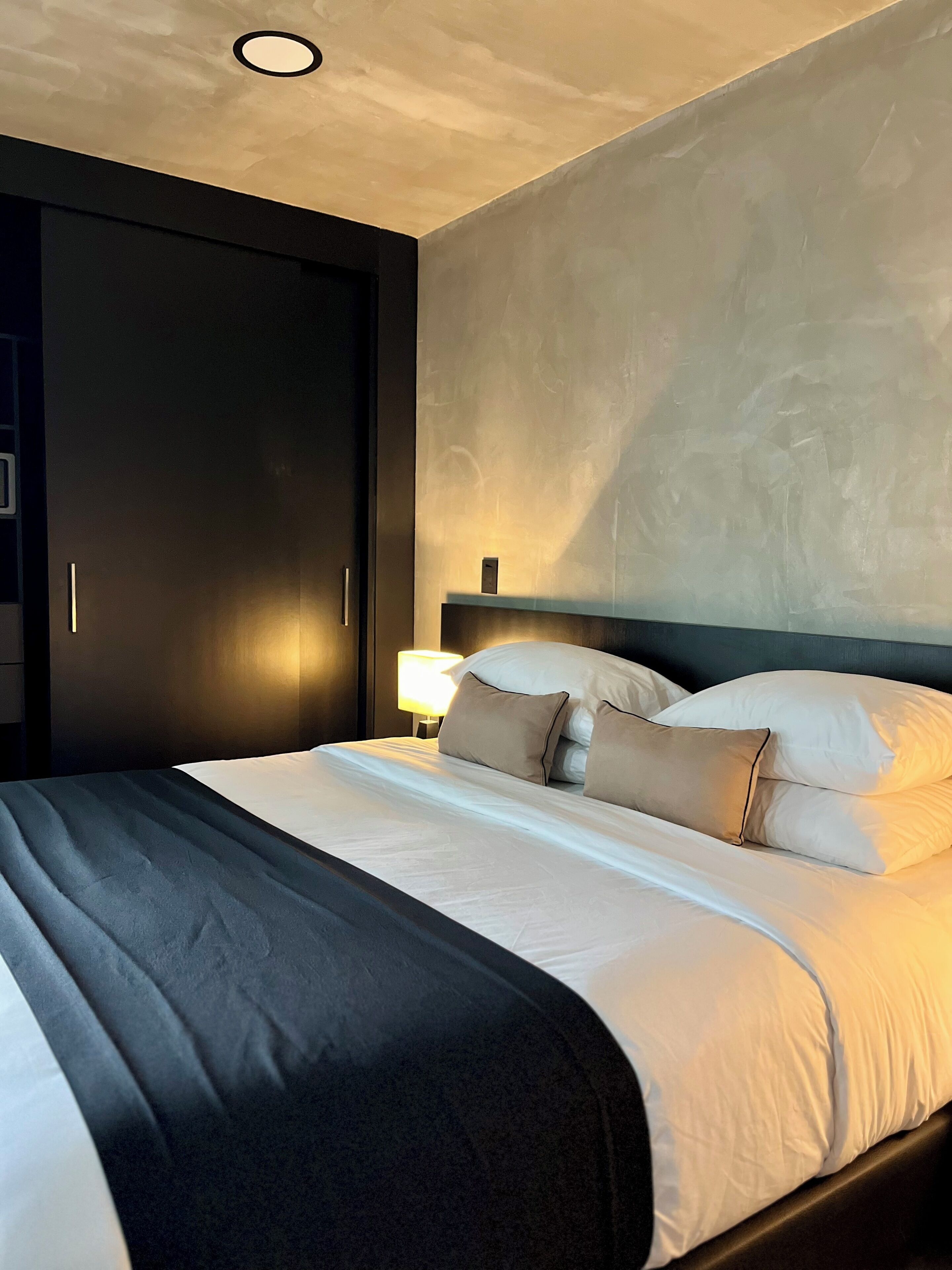 Un lit bien fait, avec du linge de lit blanc et deux oreillers beige, une lampe de chevet et une armoire de couleur sombre.