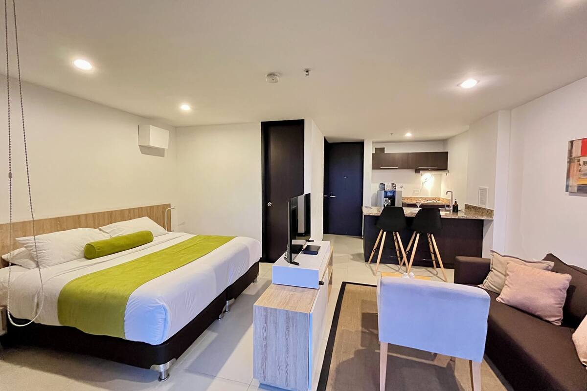 A modern hotel room with a bed, a desk, a chair, a sofa, and a kitchenette.