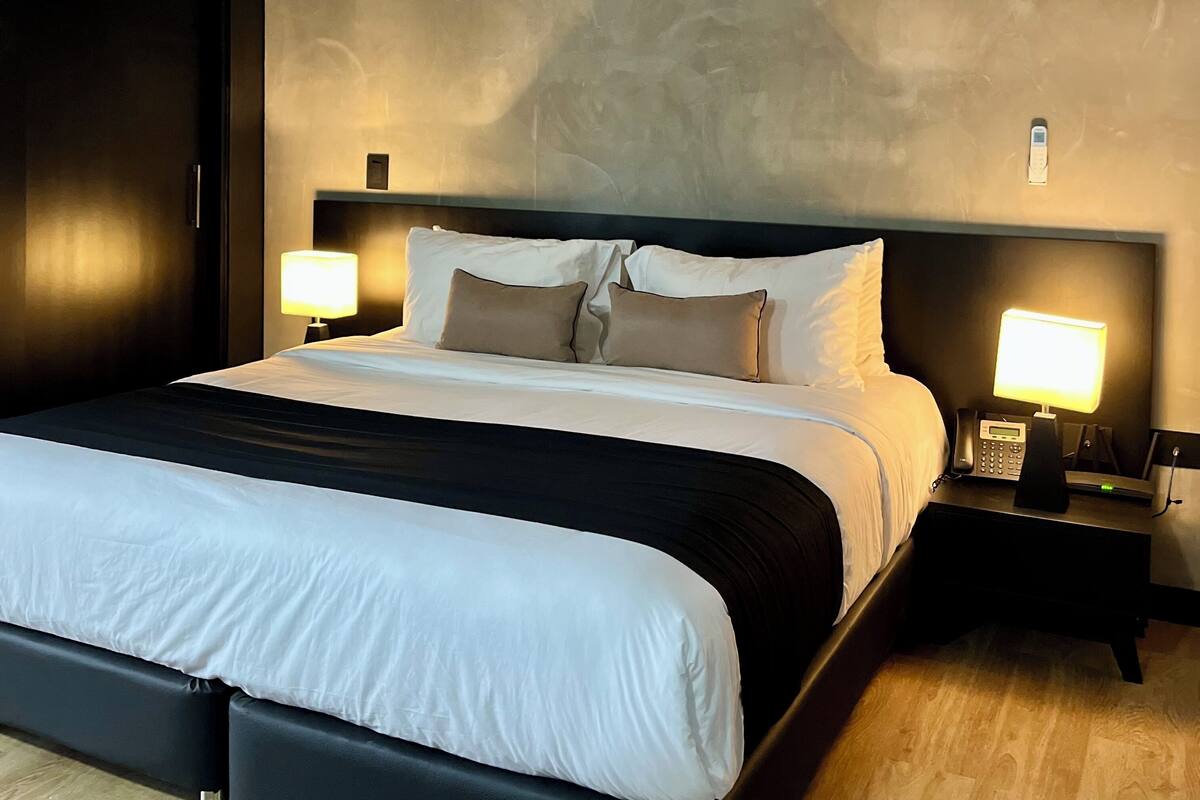 A modern hotel room with a large bed, two bedside tables with lamps, an air conditioning unit, and a dark-colored wardrobe.