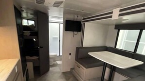A compact RV interior with a kitchenette, a seating area with a table, and a TV mounted on the wall.