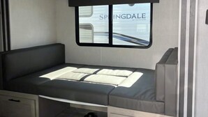 A compact RV interior with a bed, storage drawers, and a window with a view of a building labeled "SPRINGDALE."