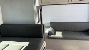 A modern RV interior with a black sofa, a smaller grey sofa, and a white bed with a pillow.