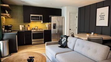 Una cocina moderna con microondas, refrigerador y estufa. En la sala de estar, hay un sofá gris y una mesa redonda de madera.