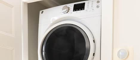 A stacked washer and dryer unit in a laundry room with a patterned tablecloth and a plastic container with items on it.