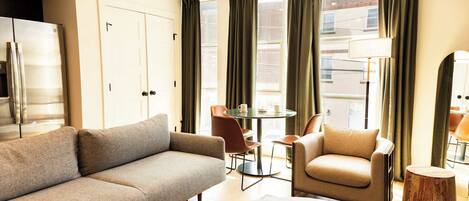 A modern hotel room with a sofa, armchair, round coffee table, and a small wooden stool.