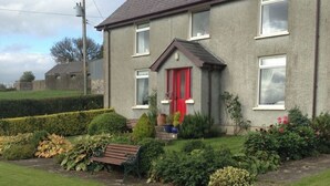 Une maison à deux étages avec une porte rouge, un petit jardin devant avec un banc et une haie.