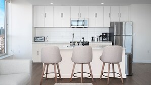 Eine moderne Küche mit weißen Schränken, einem Edelstahlkühlschrank, einer Mikrowelle und einer Kaffeemaschine. Vor der Arbeitsplatte stehen drei Barhocker.
