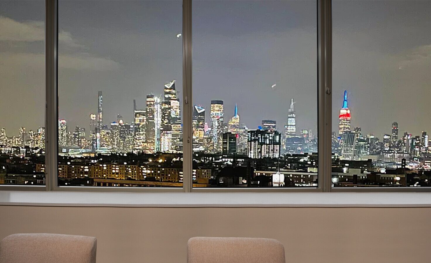Uitzicht op een stadshemelbeeld door grote ramen, met meerdere hoge gebouwen en een opvallende toren met rode en blauwe verlichting.