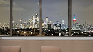 Blick auf eine Stadtlandschaft durch große Fenster, mit mehreren Hochhäusern und einem markanten Turm mit roten und blauen Lichtern.