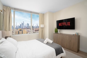 A hotel room with a large bed, a TV mounted on the wall displaying 'NETFLIX', a view of a city skyline through large windows, and a wooden console table with a plant and a tissue box.