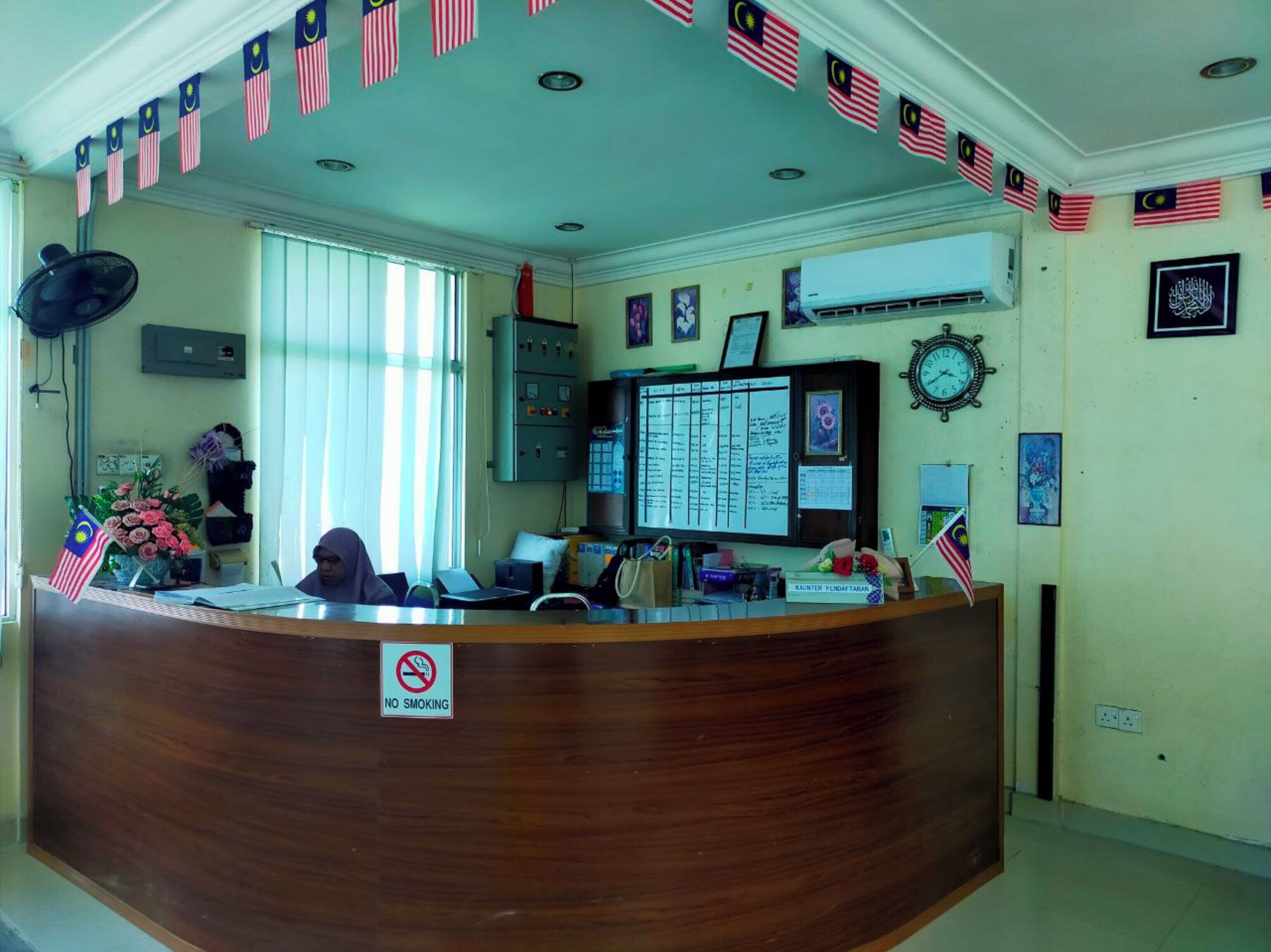 A reception desk with a no-smoking sign, surrounded by Malaysian and American flags, and a wall-mounted clock.