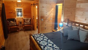 Ein gemütliches Blockhausszimmer mit Bett, zwei Sessel, einem kleinen Tisch und einem Fenster mit Jalousien.