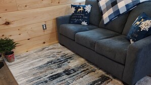 Ein gemütliches Zimmer mit grauem Sofa, gemustertem Teppich, Holzwänden und einem kleinen Topfpflanzen.