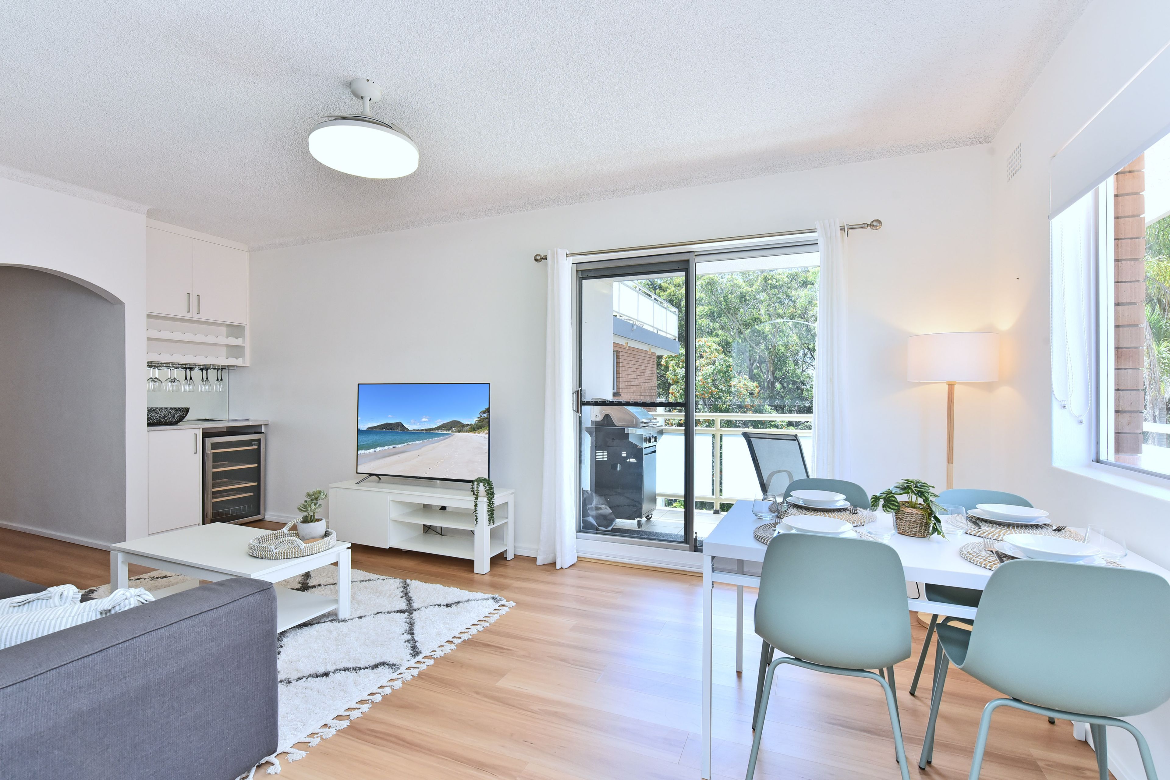 A modern living room with a flat-screen TV, a dining area with a table set for two, and a sliding glass door leading to a balcony.