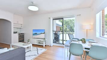 A modern living room with a flat-screen TV, a dining area with a table set for two, and a sliding glass door leading to a balcony.