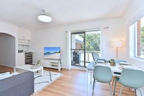 Een moderne woonkamer met een flatscreen-tv, een eethoek met een tafel gedekt voor twee personen, en een glazen schuifdeur naar een balkon.