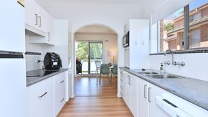 A modern kitchen with white cabinetry, a built-in microwave, and a large window overlooking a balcony. - Shoal Bay Sands - 2-Bedroom (Nelson Bay)