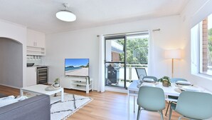 A modern living room with a flat-screen TV, a dining area with a table and chairs, and a large sliding glass door leading to a balcony. - Shoal Bay Sands - 2-Bedroom (Nelson Bay)