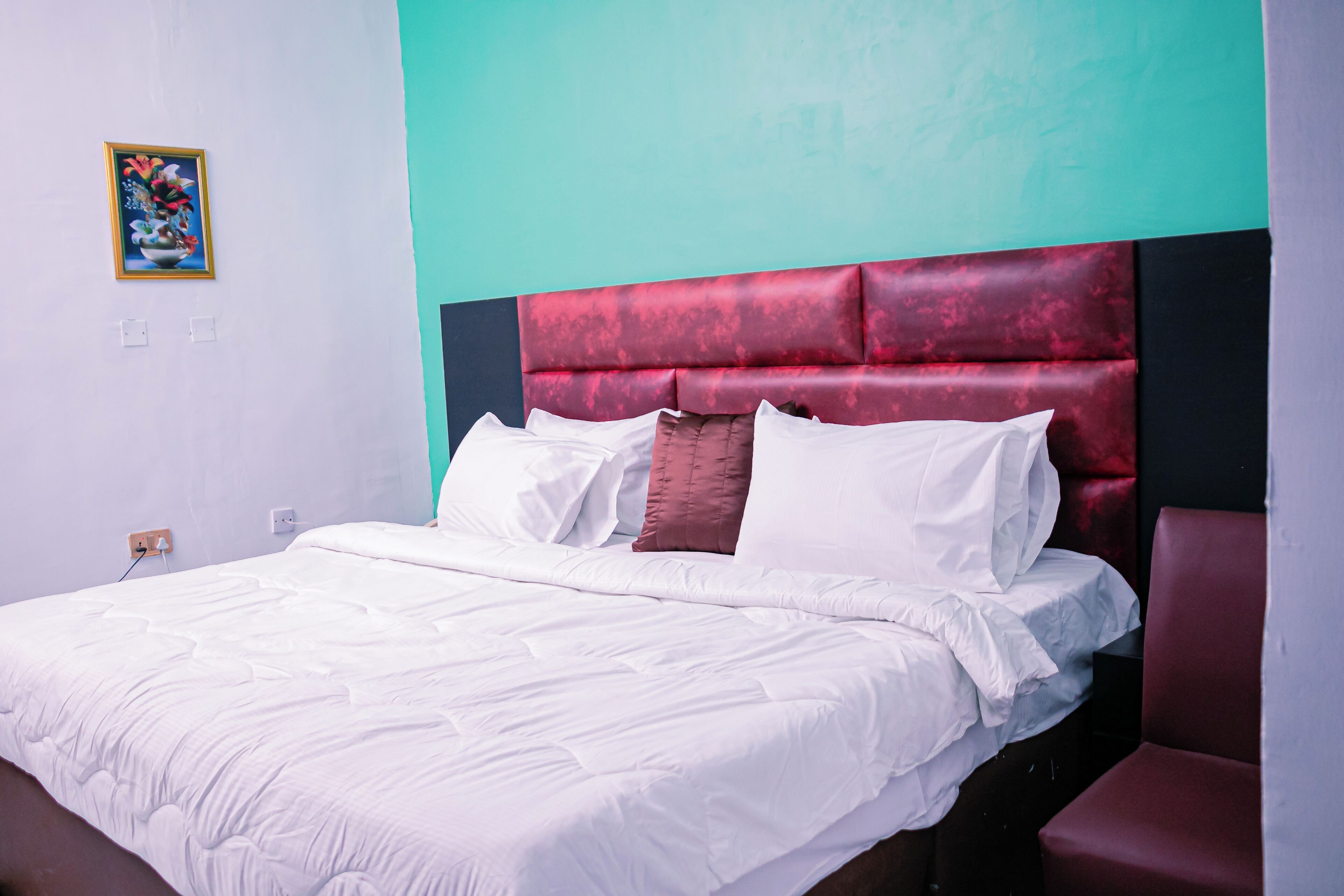 Una cama bien hecha con sábanas blancas, cabecera tapizada en rojo y una pared de acento color verde azulado.